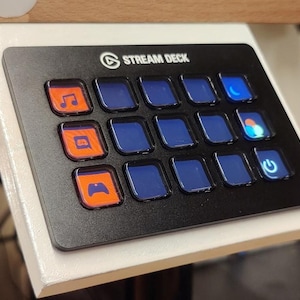 May include: Black Stream Deck with 15 illuminated buttons. The buttons are square with blue or orange backgrounds and various icons. The top of the device reads "STREAM DECK" in white text. The device is mounted on a white surface.