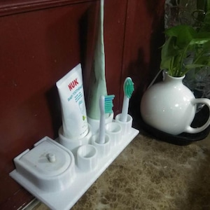 May include: A white 3D-printed toothbrush holder with multiple compartments for toothbrushes, toothpaste, and a soap dish. The holder is on a marble-patterned surface, with a tube of NUK baby toothpaste and two toothbrushes visible.