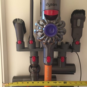 May include: A wall-mounted storage rack for a Dyson vacuum cleaner with a variety of attachments, including a crevice tool, a dusting brush, and a mini-motorized head. The rack is made of gray plastic and has red accents. The vacuum cleaner is a Dyson V8 Absolute and is orange and gray. A measuring tape is positioned below the rack, showing a measurement of 15 inches.