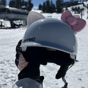 May include: A light blue ski helmet with a pink bow and white ears attached. The helmet has the word "D-TEC" printed on the front. The background shows a snowy ski slope and a ski lift.