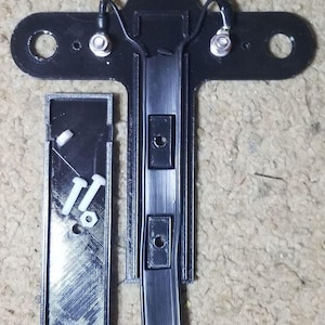 May include: Black plastic component with a cross-shaped base, two circular holes, and attached wires. A separate rectangular piece with screws and a matching hole is next to it. The item is likely an electronic part.