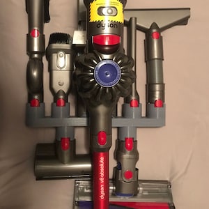 May include: A gray and red Dyson V8 Absolute cordless vacuum cleaner with a variety of attachments, including a crevice tool, a dusting brush, and a combination tool. The vacuum cleaner is mounted on a wall-mounted charging station.