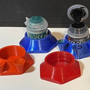 Könnte beinhalten: Drei 3D-gedruckte Farbtopfhalter, zwei blau und einer rot. Die blauen Halter haben eine kleine Vertiefung für einen Farbtopf und drei kleine Vertiefungen für Wasser oder andere Flüssigkeiten. Die roten Halter haben drei kleine Vertiefungen für Wasser oder andere Flüssigkeiten.