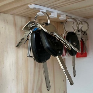 May include: A white plastic key rack with multiple hooks mounted to a wooden surface. A set of keys with silver and black keychains are hanging from the hooks.