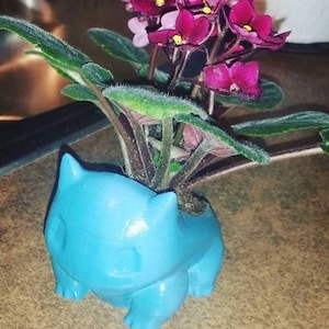 May include: A blue, 3D printed planter shaped like a Pokemon character, holding a small plant with pink flowers and green leaves.