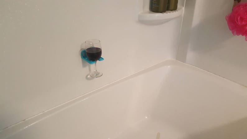 May include: A blue glass holder with a glass of red wine attached to a white tiled shower wall.