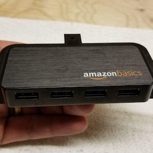 May include: Black Amazon Basics USB hub with four ports. The hub has a textured top and the Amazon Basics logo in orange. It has a rectangular shape with a small tab on top and two side tabs.