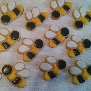 May include: A collection of ten decorated sugar cookies shaped like bees. The cookies are yellow and black with white icing accents.