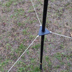 Puede incluir: Un poste de metal negro con una base triangular y tres cuerdas blancas que se extienden hacia afuera. El poste está colocado en una zona cubierta de hierba con suelo húmedo. Las cuerdas están unidas a la base con pequeños conectores blancos.