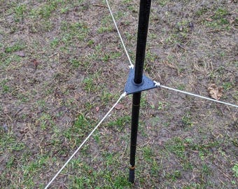 Könnte beinhalten: Ein schwarzer Metallpfahl mit dreieckiger Basis und drei weißen Seilen, die sich nach außen erstrecken. Der Pfahl steht in einem grasbewachsenen Bereich mit feuchtem Boden. Die Seile sind mit kleinen weißen Verbindern an der Basis befestigt.