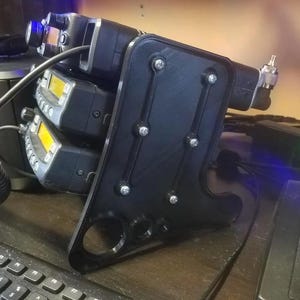 May include: Black radio mount holding two-way radios with yellow displays. The mount is attached to a black base with silver screws. A keyboard and other electronic devices are in the background.