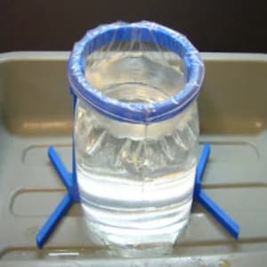 Può includere: Un barattolo di vetro trasparente con un anello di plastica blu attorno alla parte superiore, appoggiato su un supporto di plastica blu. Il barattolo è pieno d'acqua.