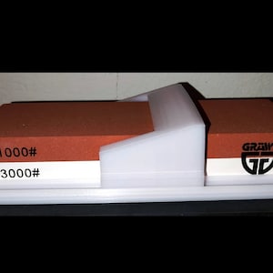 Puede incluir: Un soporte de acero para afilar de plástico blanco con dos piedras de afilar rojas y blancas. Las piedras están etiquetadas con "1000#" y "3000#" y tienen la palabra "GRAWER" impresa en ellas.