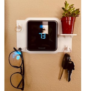 以下が含まれることがあります： 「73」を表示するHoneywellサーモスタットを備えた白い壁掛けオーガナイザー。鉢植えの多肉植物、鍵付きキーリング、メガネ用のリングが付いています。メガネは黒縁です。