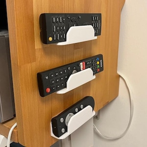 Puede incluir: Tres mandos a distancia negros se sujetan con soportes blancos montados en la pared de un mueble de madera. Los mandos tienen varios botones y están diseñados para dispositivos electrónicos.