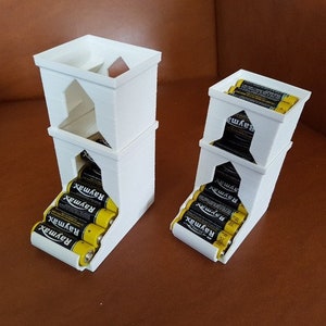 May include: Two white 3D printed battery organizers. The larger organizer is stacked three levels high and holds 6 AA batteries. The smaller organizer is stacked two levels high and holds 4 AA batteries. Both organizers have a slot for the batteries to slide into.