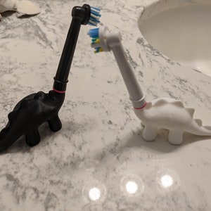 May include: Two dinosaur-shaped toothbrush holders, one black and one white, with colorful toothbrush heads. The holders are made of 3D printed plastic and have a fun, whimsical design.