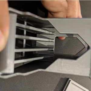 May include: A 3D printed gray plastic object with a series of parallel slots and a rectangular opening.