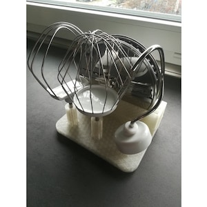 Puede incluir: Un juego de tres accesorios de color plateado para una batidora de pie. Los accesorios son un batidor, un gancho para masa y una paleta. Los accesorios están en un soporte de plástico blanco.