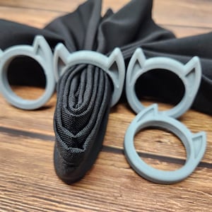 May include: Four gray cat-shaped silicone rings. The rings are designed to hold fabric together, such as a scarf or a piece of clothing.