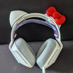 Puede incluir: Auriculares para juegos blancos con un diseño de Hello Kitty. Los auriculares tienen una diadema blanca con una banda iridiscente púrpura y azul, y dos auriculares blancos con acolchado gris. La diadema tiene un lazo rojo de Hello Kitty en la parte superior.