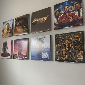 May include: Collection of vinyl record album covers displayed on a wall. The album art features diverse designs, including portraits, abstract art, and text. The covers are in various colors, including pink, brown, and black, and are mounted with white brackets.