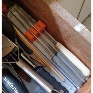 May include: A wooden drawer with a variety of kitchen utensils, including a metal grater, a wooden spoon, and a white and gray kitchen tool. The drawer is lined with a gray and white patterned liner. There are four orange rubber stoppers in the drawer.