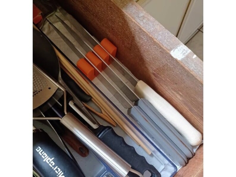 May include: A wooden drawer with a variety of kitchen utensils, including a metal grater, a wooden spoon, and a white and gray kitchen tool. The drawer is lined with a gray and white patterned liner. There are four orange rubber stoppers in the drawer.