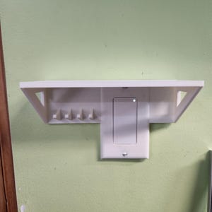 Könnte beinhalten: Ein weißes, an der Wand montiertes Regal mit integriertem Lichtschalter und vier kleinen Haken. Das Regal hat ein schlichtes, modernes Design mit dreieckigen Stützen. Der Lichtschalter ist rechteckig und hat eine kleine Kontrollleuchte.