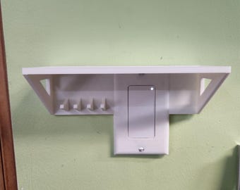 Puede incluir: Una estantería blanca montada en la pared con un interruptor de luz integrado y cuatro pequeños ganchos. La estantería tiene un diseño simple y moderno con soportes triangulares. El interruptor de la luz es rectangular y tiene una pequeña luz indicadora.