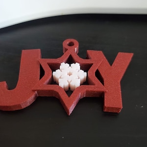 Könnte beinhalten: Roter Holzschmuck mit dem ausgeschnittenen Wort "JOY" und einem weißen Schneeflocke in der Mitte.