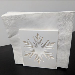 May include: White napkin holder with a snowflake design. The holder is made of plastic and has a square base.
