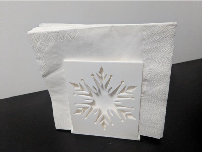 May include: White napkin holder with a snowflake design. The holder is made of plastic and has a square base.