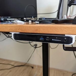Puede incluir: Un concentrador USB en blanco y negro está montado debajo de un escritorio de madera. El concentrador tiene múltiples puertos, incluyendo USB, tarjeta SD y audio. El escritorio es de madera clara, con varios cables visibles.