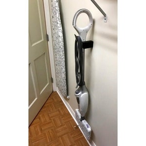 May include: A white and gray steam mop with a black handle is mounted on a wall with a black bracket. The mop is hanging vertically with the cleaning head at the bottom. The mop is in a bathroom or laundry room.