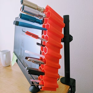 Op de afbeelding: Een feloranje zeshoekige bureau-organizer met een zwarte monitorarm die aan een bureau is bevestigd. De organizer bevat verschillende gereedschappen, waaronder schroevendraaiers, pennen en potloden.
