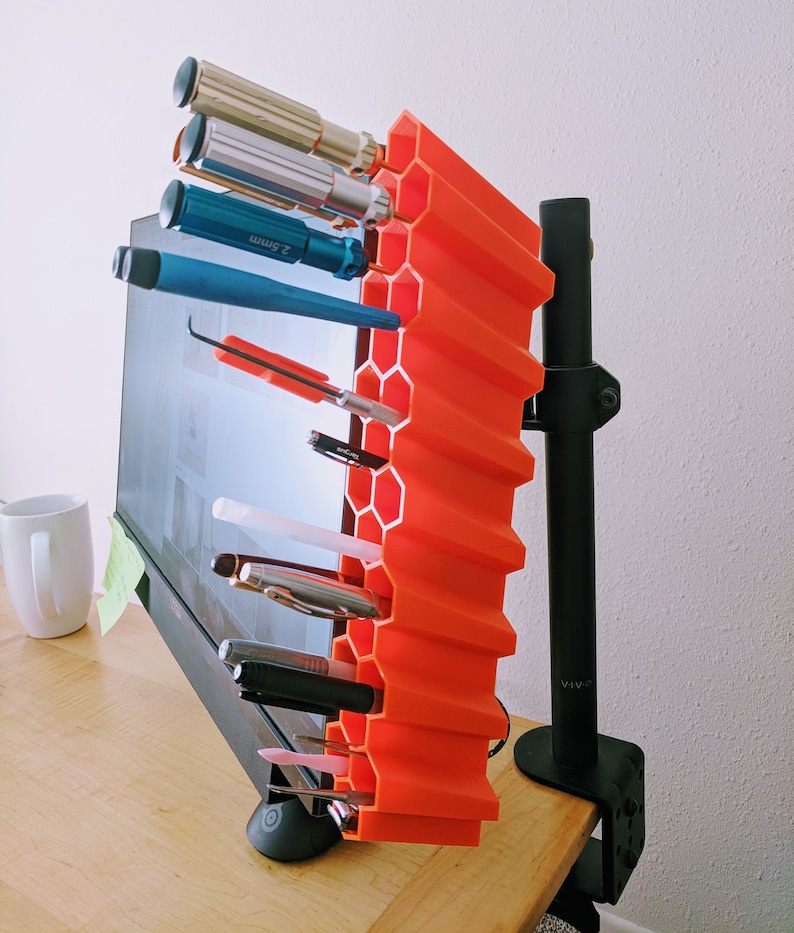 Op de afbeelding: Een feloranje zeshoekige bureau-organizer met een zwarte monitorarm die aan een bureau is bevestigd. De organizer bevat verschillende gereedschappen, waaronder schroevendraaiers, pennen en potloden.