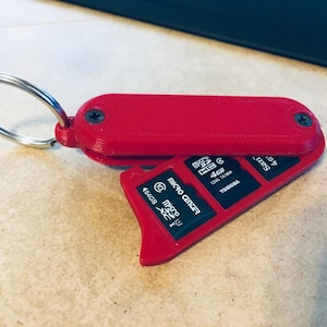 Puede incluir: Un estuche rojo para llavero impreso en 3D que contiene varias tarjetas micro SD. La funda es rectangular con bordes redondeados y asegurada con dos tornillos. Se adjunta un llavero plateado. Las tarjetas micro SD son negras con texto blanco y rojo.