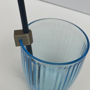 Puede incluir: Un soporte para pajitas impreso en 3D de color plateado sujeto al borde de un vaso de vidrio azul claro con líneas verticales.