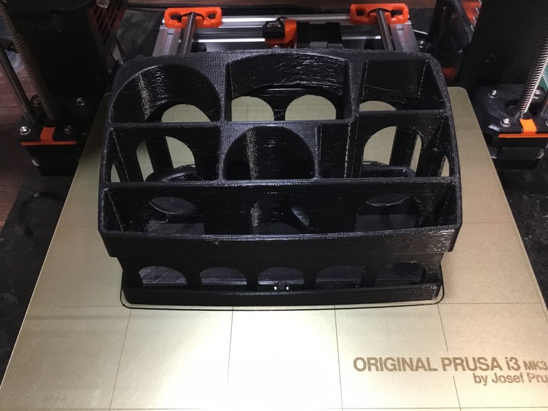 May include: A black 3D printed object with a grid-like design. The object is sitting on a beige surface with the text "ORIGINAL PRUSA i3 MK3 by Josef Prusa" printed on it.