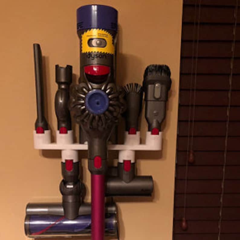 May include: A white wall-mounted storage rack for a Dyson vacuum cleaner and its attachments. The rack has five slots for holding the vacuum cleaner, a crevice tool, a dusting brush, a combination tool, and a mini-motorized head. The vacuum cleaner is purple and gray, and the attachments are all gray.
