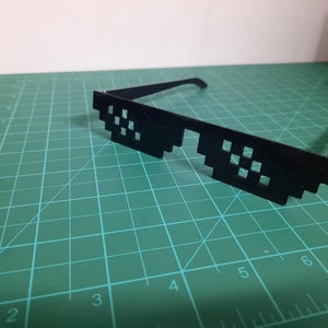 May include: Black plastic novelty sunglasses with a pixelated design. The sunglasses are on a green cutting mat.
