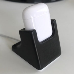 May include: A black plastic charging stand for a white wireless charging case. The stand has a rounded base and a rectangular opening for the case.