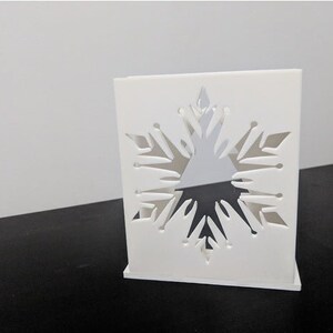 May include: White square box with a snowflake cutout design. The snowflake is intricate and detailed, with a three-dimensional effect.