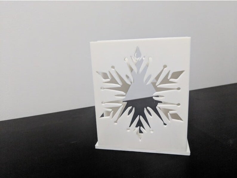 May include: White square box with a snowflake cutout design. The snowflake is intricate and detailed, with a three-dimensional effect.