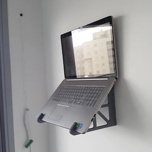 Peut inclure: Un ordinateur portable Dell argenté monté sur un support mural gris. L'ordinateur portable est ouvert, reflétant un bâtiment. Le support est incliné pour maintenir l'ordinateur portable contre le mur.