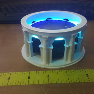 Op de afbeelding: Een witte 3D-geprinte standaard met kolommen en bogen, ontworpen om een ​​slimme speaker te houden. De standaard heeft een blauw licht dat van de basis uitstraalt.
