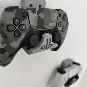 Puede incluir: Un mando de videojuegos con estampado de camuflaje montado en una pared con un soporte gris. El mando es negro y gris con un diseño de camuflaje. Un segundo mando blanco también está montado en la pared. Los soportes son grises.