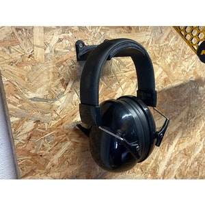 May include: Black noise-canceling headphones hanging on a wall-mounted headphone holder. The holder is made of black plastic and has a simple design.