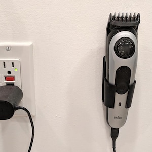 May include: A silver and black Braun beard trimmer with a black charging cable is mounted on a white wall. A black charging adapter is plugged into a wall socket.